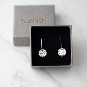 May include: A pair of silver hammered disc earrings displayed in a gray box with the word "burnish" embossed on the lid. The earrings have a textured surface and are suspended from thin silver wires.