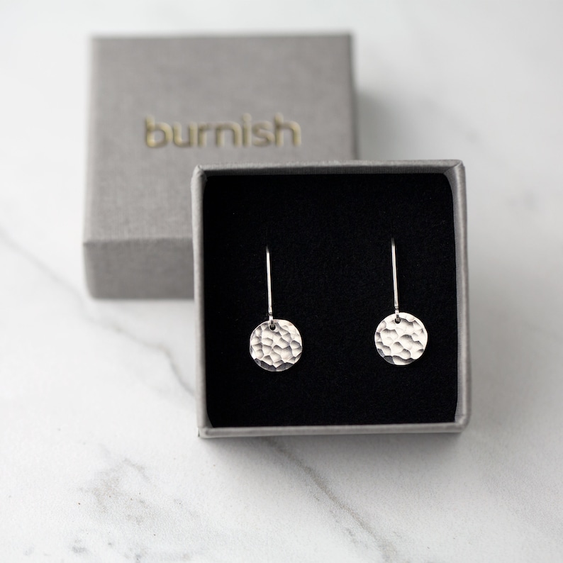 May include: A pair of silver hammered disc earrings displayed in a gray box with the word "burnish" embossed on the lid. The earrings have a textured surface and are suspended from thin silver wires.