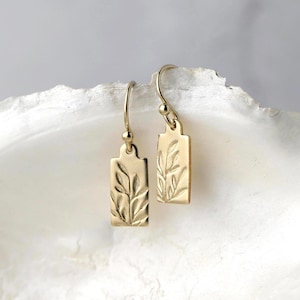 May include: A pair of gold-colored rectangular earrings with a leaf design. The earrings have a hook closure and a small ball detail. The leaf design is etched into the metal, creating a textured appearance. The earrings are displayed on a white surface.