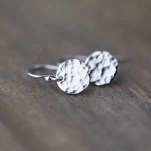 May include: A pair of silver earrings with hammered, textured circular pendants. The earrings have a simple hook design and are displayed on a wooden surface. The pendants have a textured, hammered appearance.