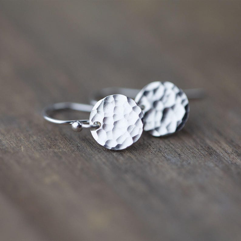 May include: A pair of silver earrings with hammered, textured circular pendants. The earrings have a simple hook design and are displayed on a wooden surface. The pendants have a textured, hammered appearance.