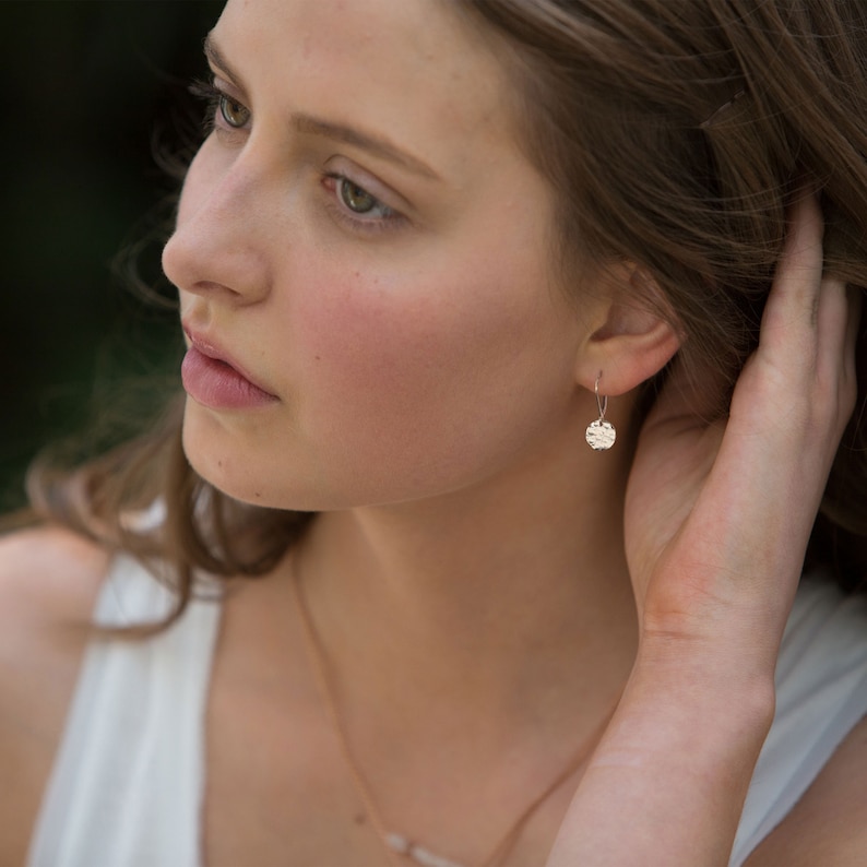 May include: A close-up of a person wearing a pair of small, silver drop earrings. Each earring features a textured, circular design. The person is wearing a white top and a delicate necklace. The background is blurred, with natural light.
