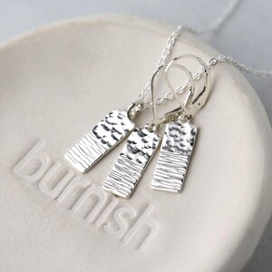 May include: A silver jewelry set featuring a necklace and two pairs of earrings. The rectangular pendants have textured surfaces with a hammered and lined design. The jewelry is displayed on a white surface with the word "burnish" embossed.