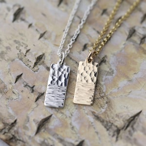 May include: Two pendant necklaces with rectangular pendants. One is silver, the other gold. Each pendant has a textured upper half and a smooth lower half, suspended from delicate chains. The background is a light-colored wood surface.
