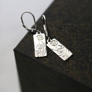 May include: A pair of silver rectangular drop earrings. Each earring features an engraved floral design with stems and leaves. The earrings have a lever back closure. The earrings are displayed on a dark surface.