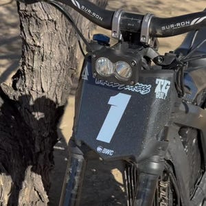 May include: Close-up of a black electric dirt bike with the number 1 in white on the front. The handlebars have the brand name SUR-RON. The bike has a dual headlight and is parked near a tree.