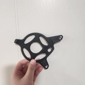 Puede incluir: Un objeto negro, circular, impreso en 3D con múltiples recortes ovalados y circulares. El objeto tiene dos pequeños agujeros en el lado derecho y una pequeña extensión en el lado izquierdo. El objeto se sostiene contra un fondo blanco.