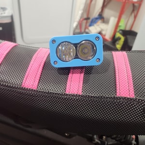 May include: A blue LED light with two circular lights. The light is mounted on a black surface with pink straps. The light has a rectangular shape with rounded corners and is secured with four screws.