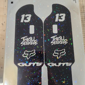 Puede incluir: Dos calcomanías negras y brillantes con el número 13 en blanco. Las calcomanías tienen las palabras "THRILL SEEKERS" y "GUTS" en blanco, junto con un logotipo de zorro blanco. La parte inferior de las calcomanías dice "BWCUSTOMS".