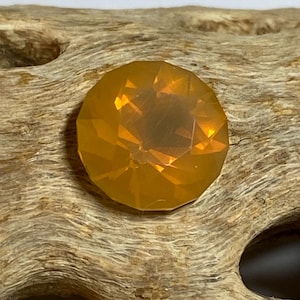 May include: A faceted, round, amber-colored gemstone. The gem has a brilliant cut with multiple facets that catch the light, creating a sparkling effect. The gemstone is set against a textured, light brown wooden surface.