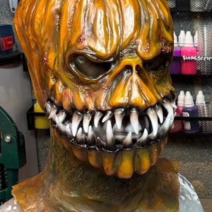 May include: A detailed, orange pumpkin mask with a menacing expression. The mask features sharp, white teeth, dark eye sockets, and a textured surface. The mask is likely for Halloween or costume use.