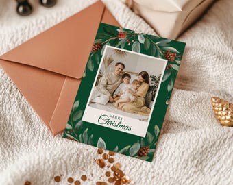 Peut inclure: Une carte de Noël avec une photo de famille, entourée d'une bordure florale verte avec des pommes de pin et des baies. La carte porte l'inscription "Merry Christmas". Une enveloppe cuivrée et des décorations dorées complètent l'ensemble.