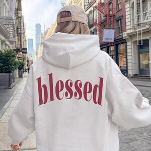 May include: A white hoodie with the word "blessed" printed in burgundy across the back. The hoodie has a hood and long sleeves. The person is wearing a tan baseball cap.