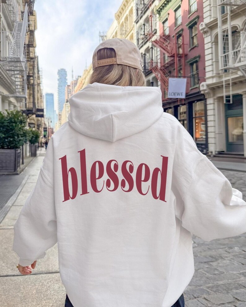 May include: A white hoodie with the word "blessed" printed in burgundy across the back. The hoodie has a hood and long sleeves. The person is wearing a tan baseball cap.