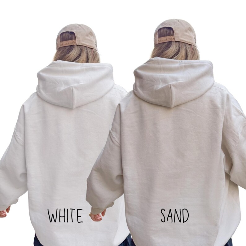 May include: Two hooded sweatshirts, one white and one sand-colored, are displayed. The word "WHITE" is printed on the back of the white hoodie, and "SAND" is printed on the back of the sand-colored hoodie. Both models are wearing a tan baseball cap.