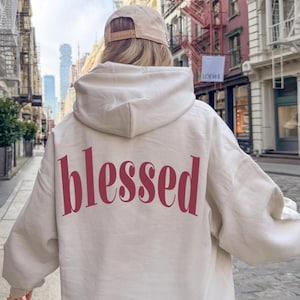 May include: A beige hoodie with the word "blessed" printed in a burgundy color across the back. The person is wearing a beige baseball cap. The background shows a city street with buildings.