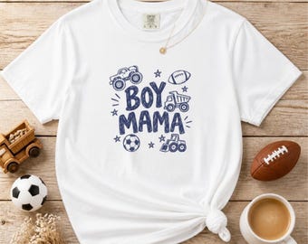 May include: A white short-sleeve t-shirt featuring a dark blue graphic print that reads BOY MAMA in the center. The design includes illustrations of a monster truck, a dump truck, a bulldozer, a football, a soccer ball, and stars.