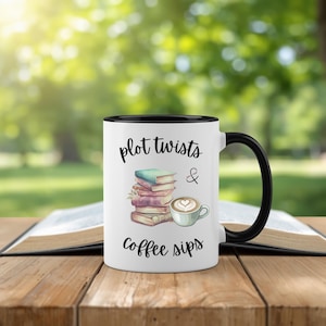 May include: White ceramic mug with a black interior and handle. The mug features the text "plot twists & coffee sips" with illustrations of books and a coffee cup. The mug is on a wooden table with an open book.