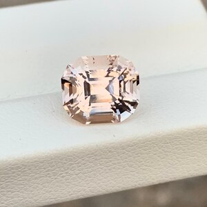 May include: A square-cut, peach-colored gemstone with a faceted surface. The gem is set on a white surface, with a blurred background of neutral tones. The gemstone's cut reflects light, creating a sparkling effect.