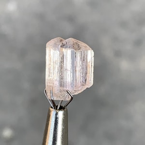 以下が含まれることがあります： 薄いピンク色の半透明の結晶が、銀色の金属製のツールで保持されています。結晶は、垂直の縞模様を持つ長方形の形状をしています。背景はぼやけたニュートラルグレーです。