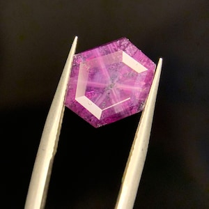 Puede incluir: Una gema hexagonal facetada, sujeta con pinzas. La gema es de un rosa vibrante con un diseño geométrico blanco. El fondo es negro, resaltando sus facetas.