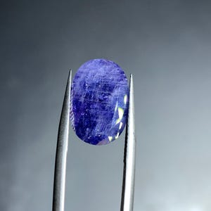May include: A faceted, oval-shaped gemstone held by tweezers. The gem is a deep, saturated blue with visible internal inclusions and facets that catch the light. The background is a gradient of gray.