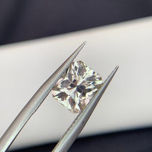 Peut inclure: Un diamant de forme carrée et de couleur claire est maintenu par une pince argentée. La pierre précieuse présente de multiples facettes qui réfléchissent la lumière, créant un effet scintillant. Le fond est un mélange de blanc et de gris foncé.