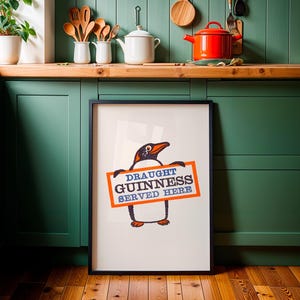May include: A framed print with a penguin illustration holding a sign that says "DRAUGHT GUINNESS SERVED HERE." The penguin is black, orange, and white. The sign is orange with blue text. The print is displayed in a kitchen setting.