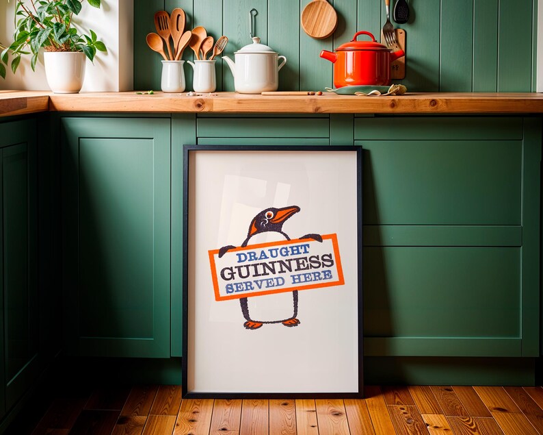 May include: A framed print with a penguin illustration holding a sign that says "DRAUGHT GUINNESS SERVED HERE." The penguin is black, orange, and white. The sign is orange with blue text. The print is displayed in a kitchen setting.