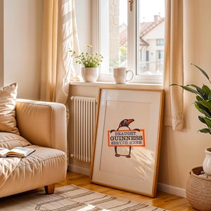May include: A framed print with a cartoon penguin holding a sign saying "Draught Guinness Served Here." The artwork is in a wooden frame, leaning against a wall. The room has a neutral colour scheme, with a sofa and a potted plant near a window.