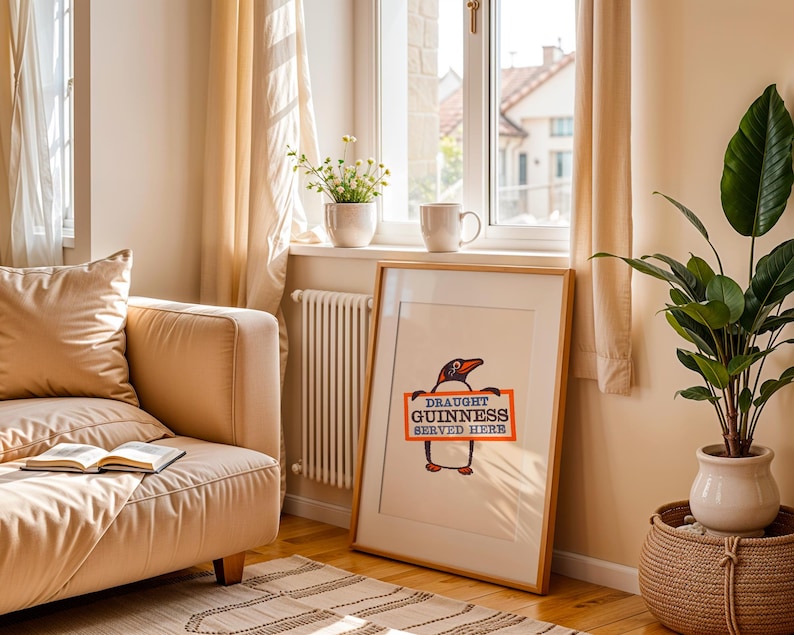 May include: A framed print with a cartoon penguin holding a sign saying "Draught Guinness Served Here." The artwork is in a wooden frame, leaning against a wall. The room has a neutral colour scheme, with a sofa and a potted plant near a window.