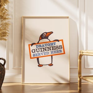 May include: A framed print with a penguin illustration holding a sign. The sign reads "DRAUGHT GUINNESS SERVED HERE" in blue and orange. The penguin is black and white with an orange beak and feet. The print is in a wooden frame.
