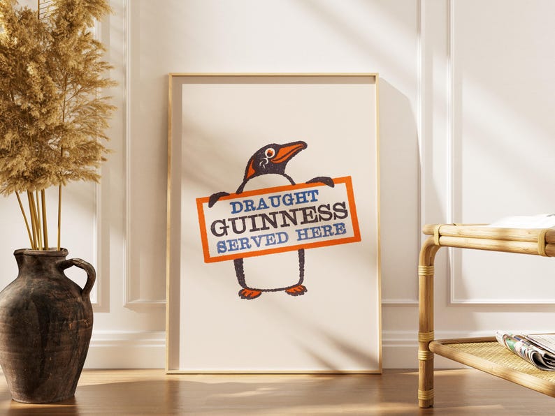 May include: A framed print with a penguin illustration holding a sign. The sign reads "DRAUGHT GUINNESS SERVED HERE" in blue and orange. The penguin is black and white with an orange beak and feet. The print is in a wooden frame.