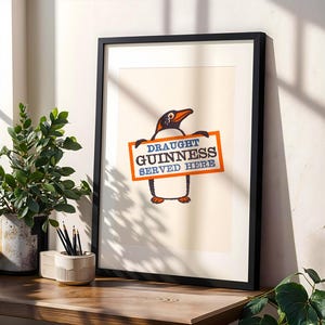 May include: Framed print of a penguin holding a sign saying "Draught Guinness Served Here." The penguin is black and white with an orange beak and feet. The sign is orange with blue and white text. The print is in a black frame, suitable for a pub or home bar.