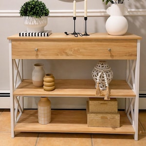 May include: A light wood console table with white metal supports, featuring a drawer and two shelves. The top shelf displays a potted plant, candles, and a vase with flowers. Decorative vases and boxes adorn the lower shelves, creating a stylish display.