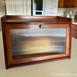 May include: A wooden bread box with a dark finish. The box has a rectangular shape with a flat top and a door with a ribbed glass panel. A small, round knob is centered on the door. The bread box is designed to sit on a countertop.