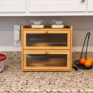 May include: A two-tiered bamboo bread box with glass paneled doors. The doors have a ribbed texture. Two small glass containers with white contents sit on top. The bread box is on a granite countertop.