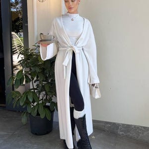 Könnte beinhalten: Eine lange, weiße, vorne offene Strickjacke mit Bindegürtel, getragen über einem weißen Oberteil mit Stehkragen und einer schwarzen Leggings. Das Outfit umfasst schwarze, kniehohe Schnürstiefel mit weißen Strickstulpen und eine kleine weiße Handtasche.