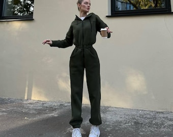 May include: An olive green hooded jumpsuit featuring a front zipper closure, long sleeves, and an elasticized waist. The garment has two side pockets and cuffed ankles, paired with white athletic sneakers and a disposable coffee cup.