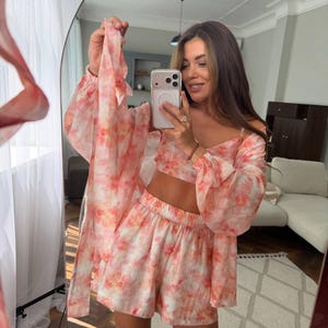 May include: A three-piece set featuring a cropped camisole top, high-waisted shorts with an elastic waistband, and a long-sleeved, open-front duster cardigan. The garments are made of a lightweight, sheer fabric with a pink and orange tie-dye pattern.