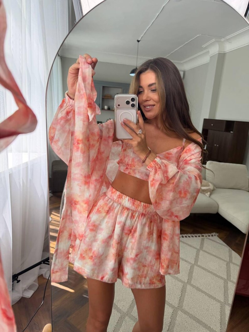 May include: A three-piece set featuring a cropped camisole top, high-waisted shorts with an elastic waistband, and a long-sleeved, open-front duster cardigan. The garments are made of a lightweight, sheer fabric with a pink and orange tie-dye pattern.