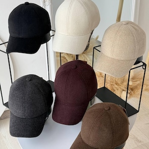 May include: A collection of six baseball caps in various solid colors including black, cream, beige, dark gray, deep burgundy, and brown. Each cap features a structured six-panel design with a curved brim and a small button at the top center.