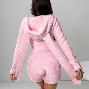 May include: A light pink, fluffy onesie with a hood and long sleeves. The onesie has attached bunny ears on the hood and a small, round tail on the back. The outfit is a soft, textured material.