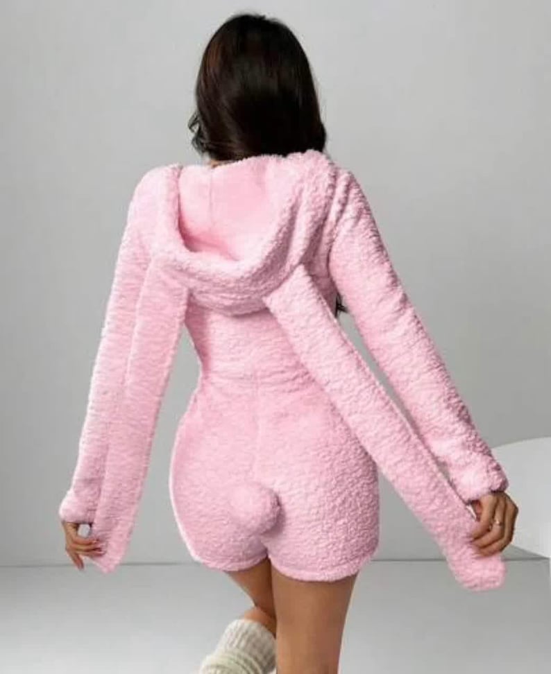 May include: A light pink, fluffy onesie with a hood and long sleeves. The onesie has attached bunny ears on the hood and a small, round tail on the back. The outfit is a soft, textured material.