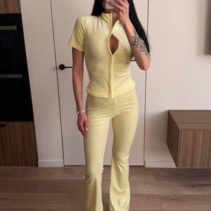 Puede incluir: Ch&aacute;ndal de terciopelo amarillo claro, compuesto por una parte superior con cremallera y pantalones acampanados. La parte superior tiene mangas cortas y cuello alto. Los pantalones tienen pernera acampanada. El conjunto se combina con zapatillas blancas.