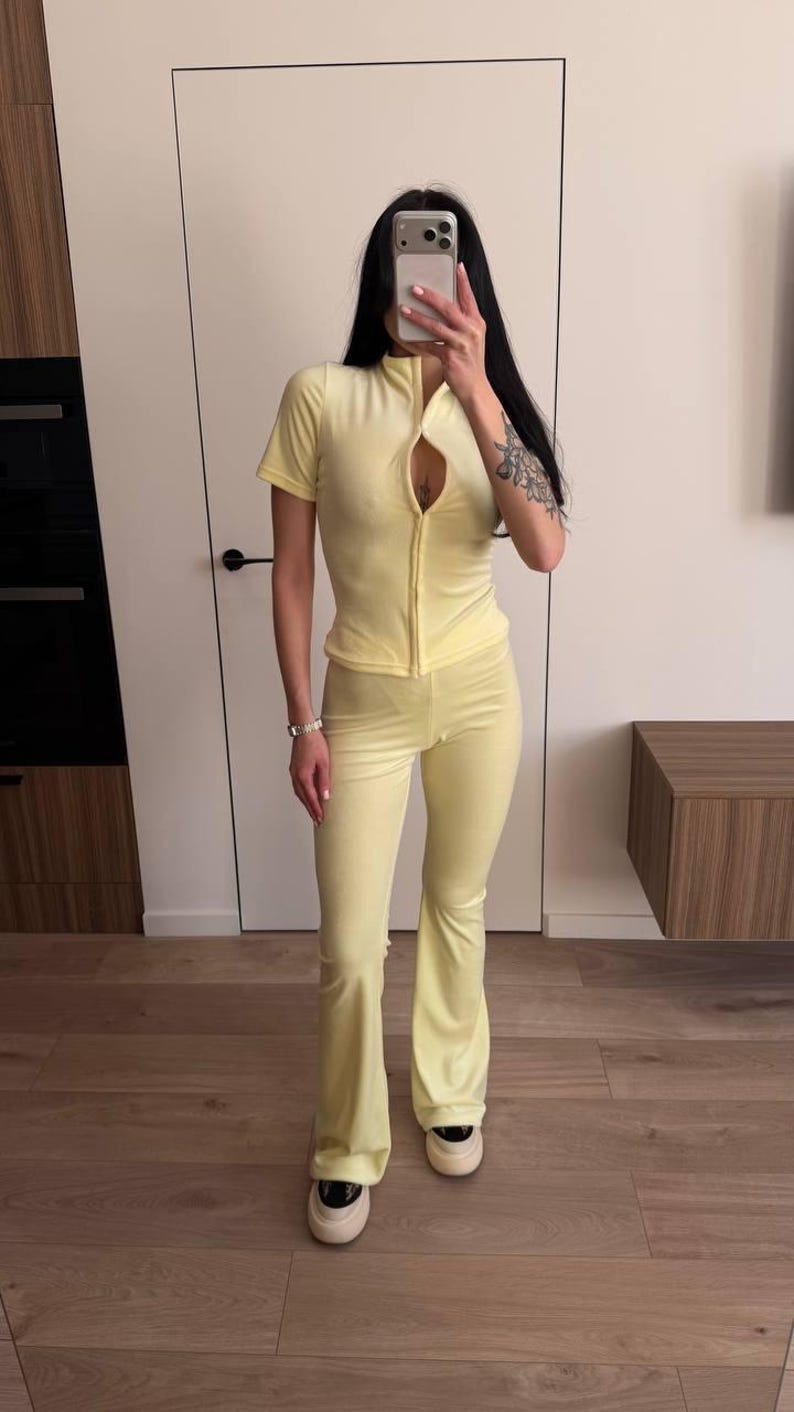 Puede incluir: Ch&aacute;ndal de terciopelo amarillo claro, compuesto por una parte superior con cremallera y pantalones acampanados. La parte superior tiene mangas cortas y cuello alto. Los pantalones tienen pernera acampanada. El conjunto se combina con zapatillas blancas.
