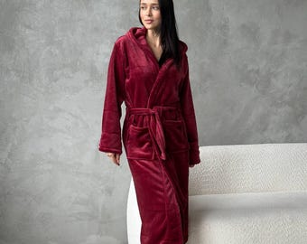 May include: A burgundy hooded robe with a matching belt and pockets. The plush robe is knee-length and worn with fluffy white slippers. The background is a neutral gray wall and a white sofa.