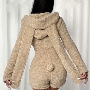 May include: A tan-colored, hooded romper with long sleeves and shorts. The romper features a fluffy, textured fabric, a hood with long, dangling ear-like extensions, and a small, round tail detail on the back.