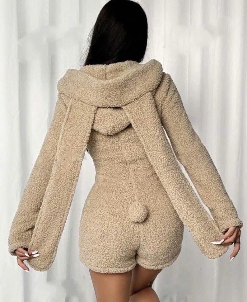 May include: A tan-colored, hooded romper with long sleeves and shorts. The romper features a fluffy, textured fabric, a hood with long, dangling ear-like extensions, and a small, round tail detail on the back.