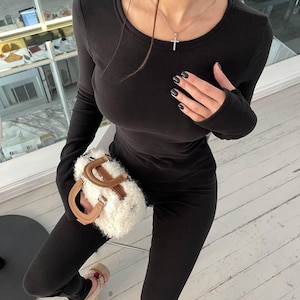 Puede incluir: Conjunto negro de top de manga larga y leggings. El atuendo se combina con un pequeño bolso blanco y esponjoso con asas marrones. Se lleva un collar con colgante de cruz plateada. La persona lleva zapatillas beige.
